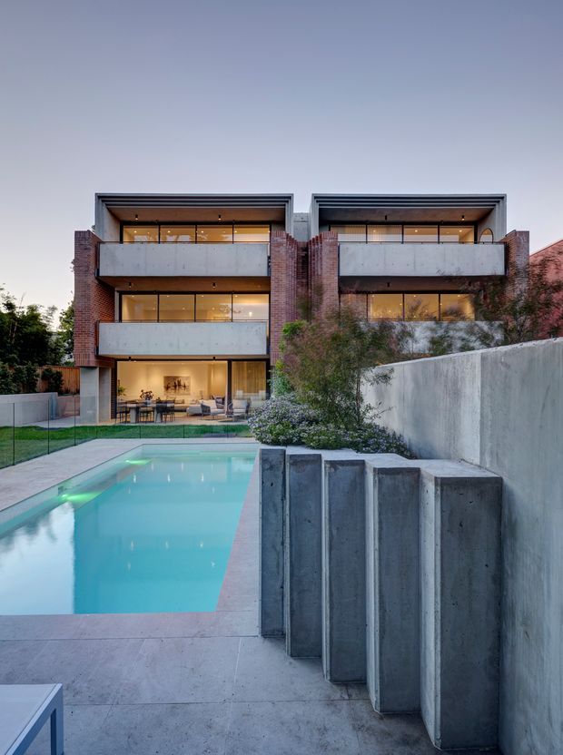 Twilight exterior view of a multi-storey contemporary residence with exposed red-brick side walls, concrete balcony slabs, dark framed glazing, and strong horizontal roofline detailing. A turquoise swimming pool and paved terrace occupy the foreground, with landscaped planting and substantial concrete retaining walls around the property.