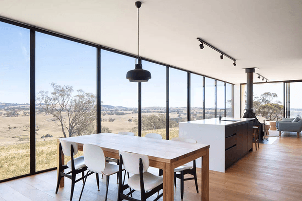 A bright open-plan dining and kitchen interior with expansive floor-to-ceiling glazing overlooking a dry rural landscape. The space features a timber dining table with white chairs, warm timber flooring, a minimalist white kitchen island with dark cabinetry, black-framed glazing, and simple contemporary pendant and track lighting.