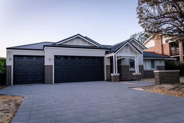 Street-facing view of a large suburban house with a white rendered facade, dark brick base and accents, multiple pitched gables, and three dark sectional garage doors. A wide grey paved driveway leads to the attached garage, with a covered entry porch and arched detailing visible at the front.