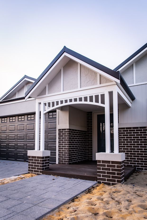Front exterior view of a newly built suburban home with prominent gabled rooflines, white rendered walls, dark blue-grey trim, black brickwork, a covered porch with decorative arched detailing, and an integrated double garage. A paved driveway and unfinished sandy landscaping are visible in the foreground.