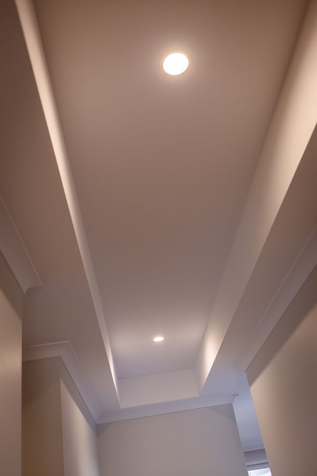 Upward-looking view of a narrow interior passage or transitional space, dominated by a recessed tray ceiling with layered plasterboard bulkheads, simple white cornice detailing, and two recessed downlights. The neutral warm-white surfaces create a clean, understated finish.