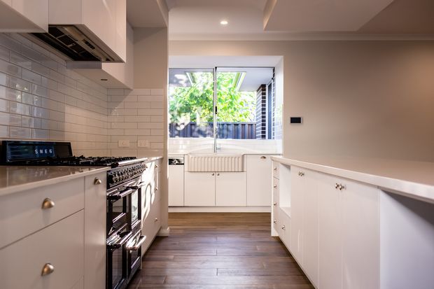Bright, refined kitchen viewed from the cooking zone toward a rear window. The room features white shaker-style cabinetry with brass knobs, a large white island, dark timber flooring, a stainless steel range cooker, glossy white subway-tile splashback, and a coffered ceiling with recessed lighting. Garden greenery is visible beyond the window.