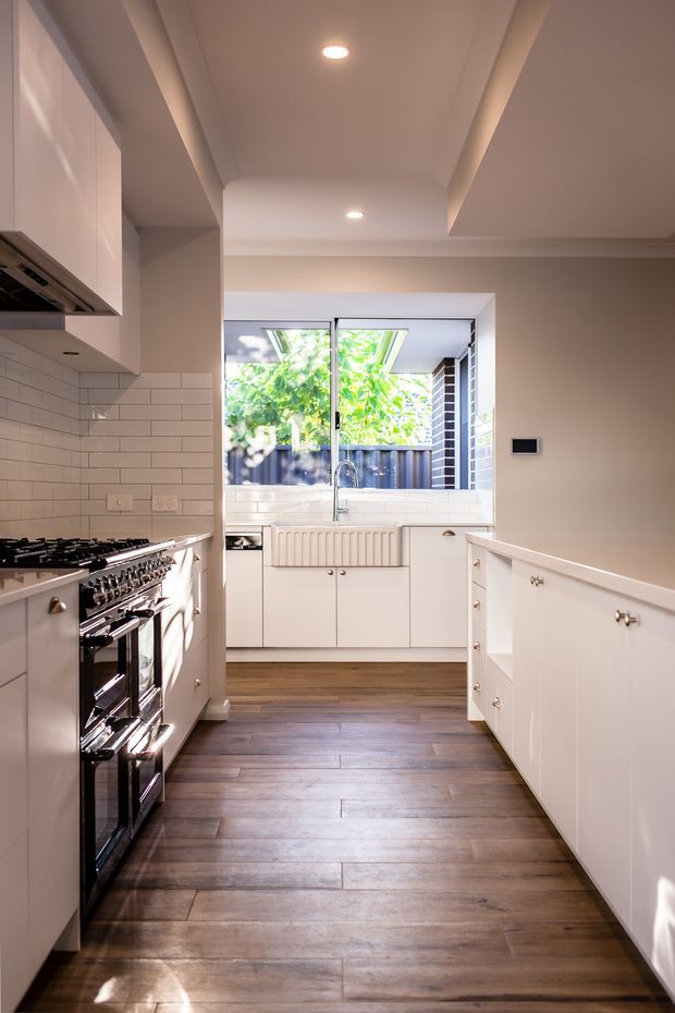 A bright, narrow galley kitchen with white cabinetry, a large freestanding stainless-steel range, white subway-tile splashback, timber-look plank flooring, and a rear sink beneath a wide window. The room has a warm neutral palette, recessed ceiling lights, and a layered ceiling bulkhead design, with greenery visible outside.