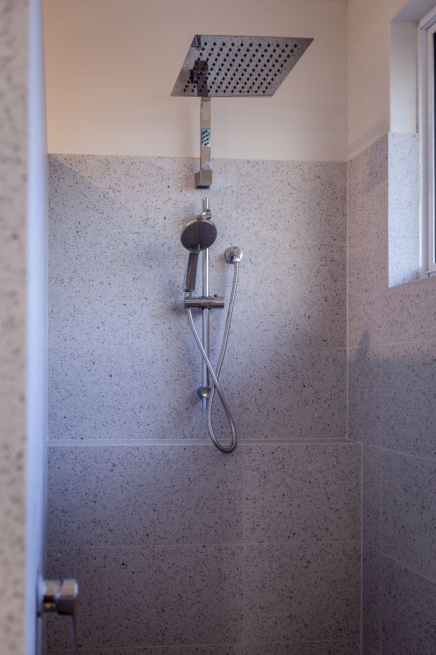 A compact modern shower enclosure viewed through a partially open door. The walls are clad in speckled light-grey stone-look tiles with matching grout, and the shower features a large square overhead rain shower, handheld shower, chrome mixer, and exposed hose. A narrow window on the right provides natural light.