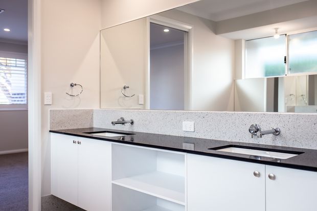 Bright contemporary bathroom vanity with a long white built-in cabinet, two integrated basins, dark benchtop, pale speckled splashback, wall mirror, chrome tapware and open shelving. A carpeted adjoining room is visible through the doorway at left.