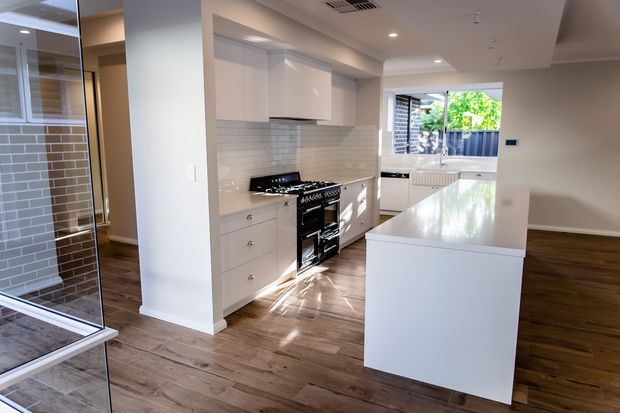 Bright contemporary kitchen with white shaker-style cabinetry, a large white island with a polished stone benchtop, stainless steel cooking range, and light grey subway-tile splashback. Warm timber flooring extends into the adjacent open-plan living area, while a glazed shower enclosure is visible at the left edge. Natural light enters through the rear window, creating a clean, airy interior.