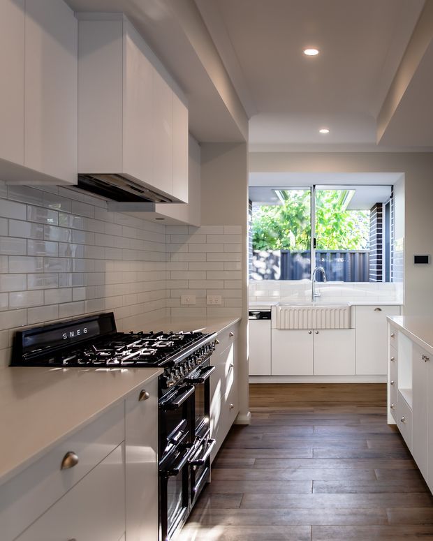 A bright, narrow galley kitchen with white shaker-style cabinetry, a large freestanding black range cooker, stainless rangehood, pale benchtops, and glossy white subway-tile splashback. Dark timber flooring runs toward a rear utility or sink area with a large window overlooking greenery and a timber privacy fence. Recessed ceiling lights and a stepped ceiling detail add depth.