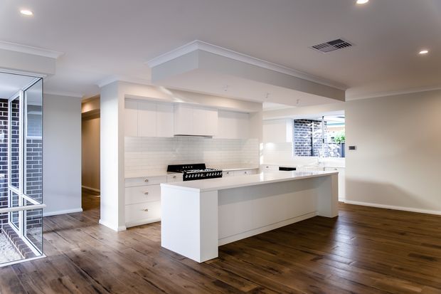 Bright open-plan kitchen and living area with white shaker-style cabinetry, a large central island, pale tiled splashback, timber flooring, recessed lighting, and white decorative cornices. A mirrored opening and brick exterior are visible at the left, while the kitchen opens into a spacious unfurnished living zone.