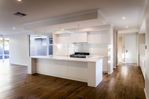 Bright open-plan kitchen with a large white island, white flat-panel cabinetry, white tiled splashback, and a freestanding cooktop. The kitchen opens to a spacious living area and hallway, with warm timber flooring, white walls, recessed lighting, and traditional-style ceiling cornices and bulkheads.