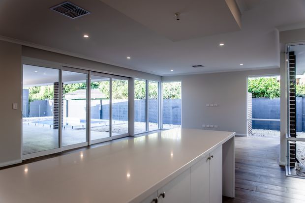 Bright contemporary open-plan kitchen with a large white island, extensive floor-to-ceiling sliding glass doors, and views to a paved outdoor entertaining area and swimming pool. The room has recessed ceiling lights, neutral painted walls, white cabinetry, and dark timber-look flooring extending toward the outdoor space.