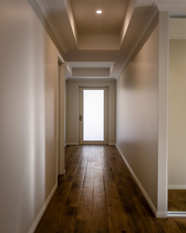 A narrow, warm-toned residential hallway with white painted walls and detailed recessed tray ceiling sections, a dark timber-look plank floor, and a frosted-glass exterior door at the far end. Open doorways and a mirrored wardrobe or closet edge are visible along the sides. The space is softly lit by a recessed ceiling light.