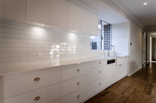 Bright, compact modern kitchen with extensive white flat-panel cabinetry, a long pale benchtop, glossy white subway-tile splashback, stainless-steel sink and tap, and integrated appliances. Warm timber flooring runs through the kitchen and adjacent hallway, with a brick feature wall visible through the window.