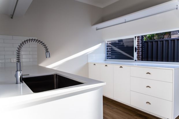 Bright contemporary kitchen featuring a deep black undermount sink with a spring-coil pull-down faucet, white cabinetry with brass cup pulls, white subway-tile splashback, and warm timber-look flooring. A wide window above the cabinetry brings in strong natural light and overlooks a fenced exterior.
