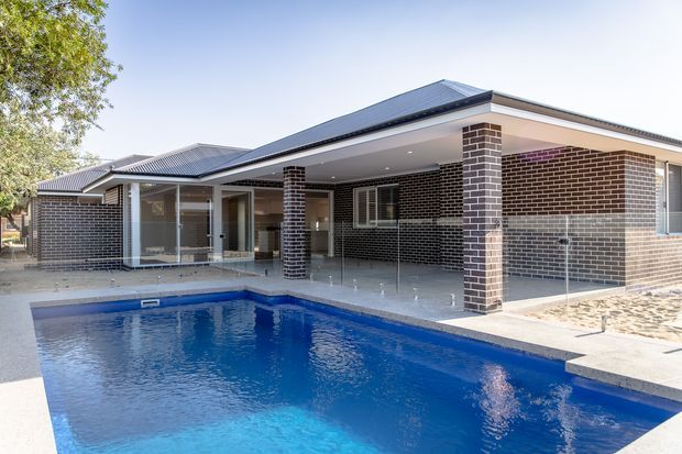 Single-storey contemporary brick home viewed from the pool, with a dark pitched metal roof, extensive covered outdoor entertaining area, glass balustrades and large glazed openings. A bright blue swimming pool dominates the foreground, while the house features dark brown brickwork and pale concrete or tiled paving.