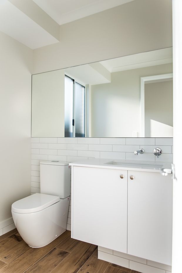 Bright compact bathroom featuring a wall-mounted white vanity with integrated basin, chrome wall-mounted tapware, a white toilet, white horizontal subway-tile splashback, large mirrored wall cabinet or panel, pale painted walls, and warm timber-look flooring. The room has a clean, contemporary and functional appearance.