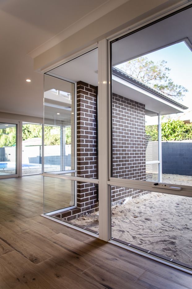 Bright modern interior featuring large white-framed glazed sliding or folding doors opening to a sandy outdoor courtyard. The room has wide-plank timber flooring, white walls and ceiling with recessed lighting, and exposed dark brickwork visible around the glazed opening. Reflections and additional glazing create a light-filled, open indoor-outdoor connection.