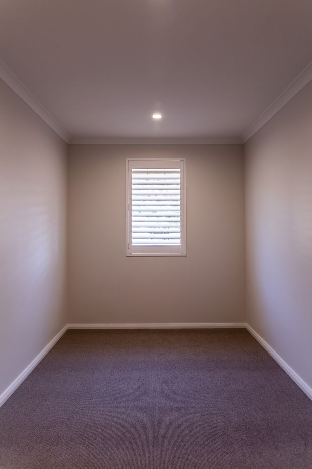A small, empty rectangular interior room with taupe painted walls, white ceiling cornices and skirting boards, mauve-grey carpet, and a single centered window fitted with white plantation shutters. A recessed ceiling light provides soft illumination.