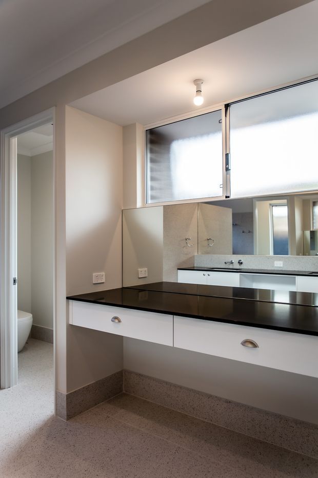 A compact, light-filled bathroom vanity area with a long black countertop, white drawers with shell pulls, a large wall mirror, and a double-basin wash area reflected in the mirror. Frosted awning windows provide natural light above the vanity. A toilet is partially visible through the open doorway on the left. The room has neutral painted walls, terrazzo-style flooring and skirting, and simple contemporary fixtures.