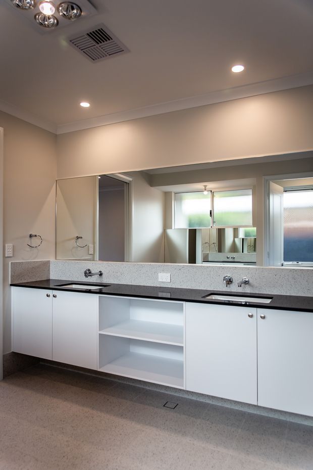 A bright, spacious bathroom with a long double vanity featuring white flat-panel cabinetry, open shelving, dark stone-look benchtops, integrated basins, chrome tapware, and a large wall-to-wall mirror. The room has pale neutral walls, recessed ceiling lights, a ceiling ventilation grille, frosted windows, and a speckled hard-surface floor.