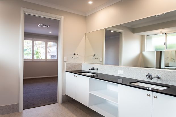 A bright, clean contemporary bathroom with a long white built-in vanity, black benchtop, two integrated basins, wall-mounted taps, a large mirrored splashback, and white cabinetry with open shelving. A carpeted bedroom or sitting room is visible through the doorway, with another mirrored or glazed area reflected in the vanity mirror.