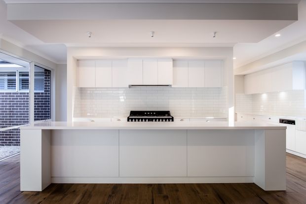 Bright contemporary white kitchen with a large central island, flat-panel white cabinetry, white subway-tile splashback, integrated rangehood and gas cooktop, and dark timber-look flooring. Glazed doors on the left open toward an exterior brick courtyard or passage.
