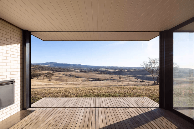A covered outdoor terrace or sunroom-like viewing space frames a broad rural landscape through large open glazing. The space features a timber-lined ceiling, timber decking, dark structural framing, and a white brick wall with a built-in fireplace or heater on the left. The expansive opening provides a strong connection to the dry, rolling countryside.