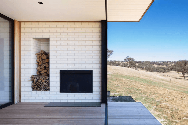 A minimalist covered outdoor living area with a white brick feature wall, recessed firewood storage, and a black built-in fireplace. Timber decking extends toward an open rural landscape, with a clean flat roof and dark-framed glazing creating a strong indoor-outdoor connection.