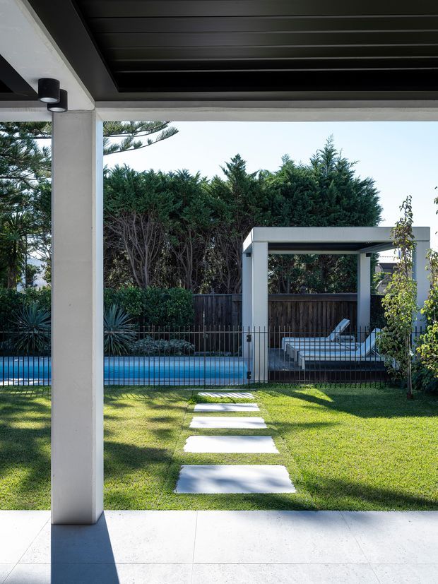 View from a covered outdoor patio across a manicured lawn toward a narrow swimming pool, stepping-stone path, and a simple white poolside pavilion with a lounge chair. The space features strong rectangular architectural lines, white structural columns, a dark slatted overhead roof, pale paving, and dense privacy planting. The image has a clean, contemporary outdoor-residential character with bright natural light and a calm garden setting.