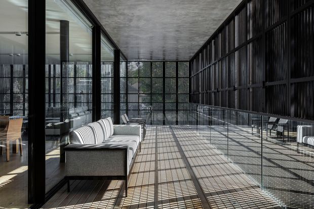 A spacious, modern covered terrace or balcony with a polished concrete ceiling, dark metal screened walls, glass balustrades, and a grey outdoor sectional sofa. Strong linear shadows from the metal screening create a graphic pattern across the pale tiled or concrete floor. Full-height glazing on the left reveals an adjacent dining and living area.