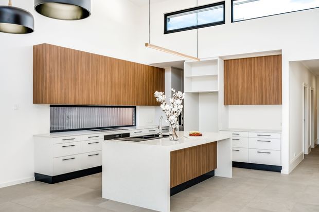 Bright contemporary kitchen with a large white island, integrated cooktop and sink, white lower cabinetry, and warm vertical-grain timber overhead cabinets and island panel. High clerestory windows, open shelving, linear pendant lighting, and dark recessed plinths create a clean, spacious interior.