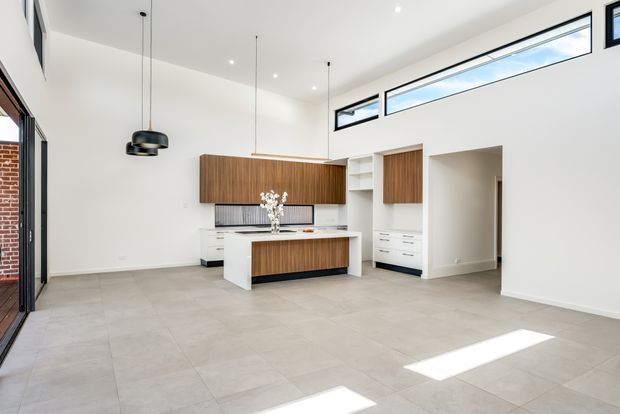 Bright, spacious contemporary kitchen and open-plan living area with a central island, warm timber cabinetry, white built-in storage, pale tiled flooring, high clerestory windows, and black pendant lighting. The room has a clean, minimalist fit-out with strong natural light and a visible brick exterior edge through the glazing.