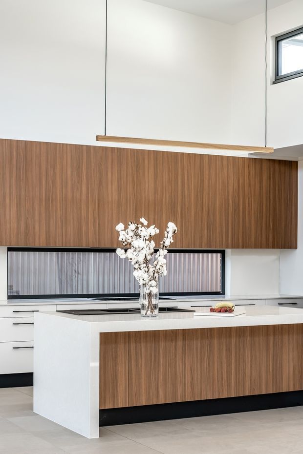 A bright, contemporary kitchen featuring a large white island with a walnut-toned timber veneer front, matching full-height cabinetry, and integrated dark appliances. A linear timber pendant hangs above the island, while a long horizontal window with ribbed screening provides natural light. The space has pale flooring, white walls, and a clean, minimalist composition.