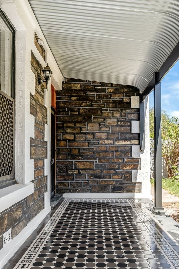 Covered exterior entry porch with a patterned geometric tiled floor, dark natural stone masonry walls, white rendered trim and columns, a curved corrugated metal canopy, and a traditional wall-mounted lantern. A security screen door and garden are visible at the sides.