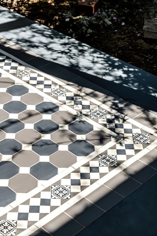 Close-up view of an outdoor tiled surface featuring a decorative geometric pattern. Large alternating dark grey and warm grey octagonal tiles are bordered by black-and-white checkerboard bands with ornate patterned square accent tiles. The surface is edged by dark blue-grey rectangular tiles and surrounded by garden planting, with strong dappled sunlight and tree shadows across the paving.