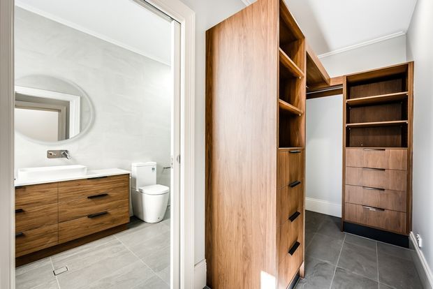 Bright interior view showing a walk-in wardrobe with extensive dark timber cabinetry, open shelving, drawers, and hanging space. Through the open doorway, a compact bathroom is visible with a timber vanity, white basin and toilet, oval mirror, pale walls, and grey tiled flooring.