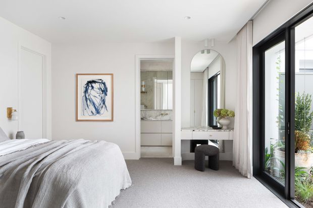 Bright, refined bedroom with a soft neutral palette, upholstered bed, full-height black-framed glazing, and sheer curtains. A compact dressing or vanity area with an arched mirror is positioned beside the bedroom, with an ensuite bathroom visible through the central opening.