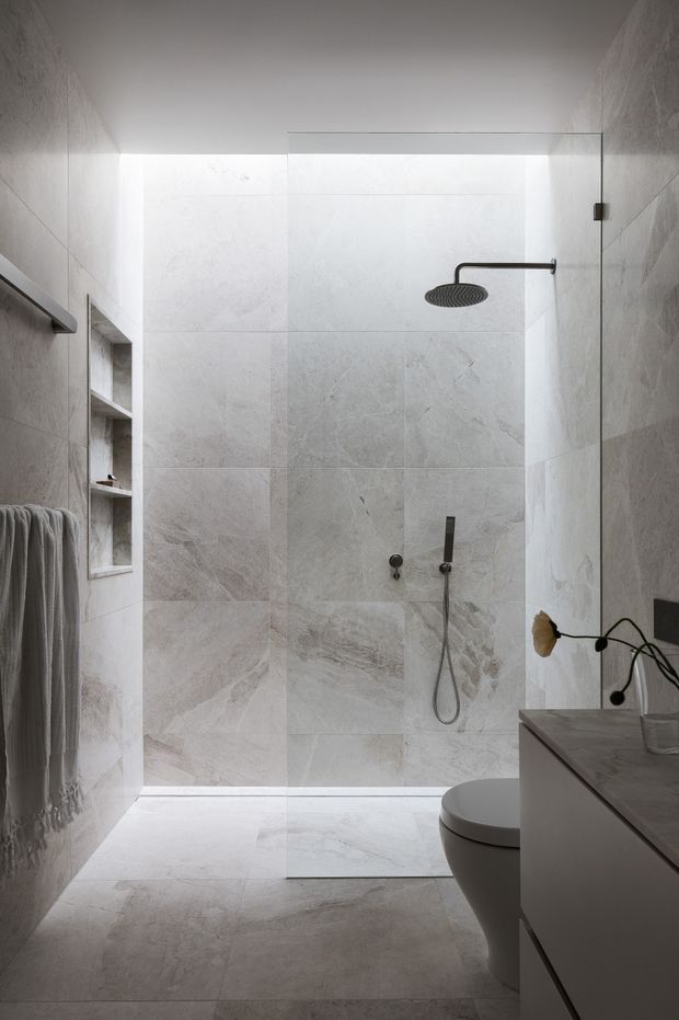 A refined contemporary bathroom with a walk-in glass shower enclosed in large-format pale stone-look tiles. The shower features a dark rainfall showerhead and hand shower, recessed shelving, a wall-mounted towel rail, and subtle integrated lighting. A floating vanity and toilet occupy the foreground, with muted grey and white finishes creating a calm, minimalist atmosphere.
