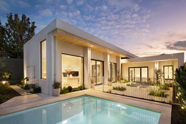 A modern single-storey residence viewed from the pool courtyard at dusk. The white rendered home features a flat roof, expansive dark-framed glazing, a warm timber-lined covered veranda, slender columns, and a landscaped pool terrace. Interior spaces are visible through the glazing, with soft architectural lighting creating a refined contemporary atmosphere.