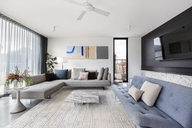 Bright contemporary living room with floor-to-ceiling sheer glazing, a large textured sectional sofa, a blue tufted sofa, patterned area rug, abstract wall art, ceiling fan, and a black media wall with a mounted television and light stone-textured feature surface.