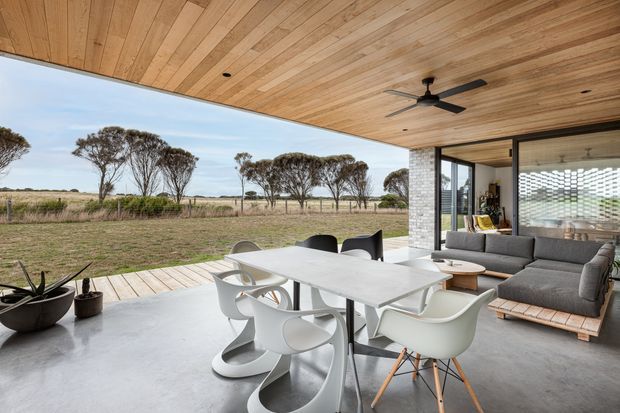Open-plan contemporary living and dining area with a large white dining table and sculptural white chairs, a grey modular sofa, and a timber-framed ceiling extending to a broad covered outdoor opening. The polished concrete floor, exposed timber ceiling, ceiling fan, glazed walls, and perforated screen create a relaxed indoor-outdoor connection overlooking a rural landscape.