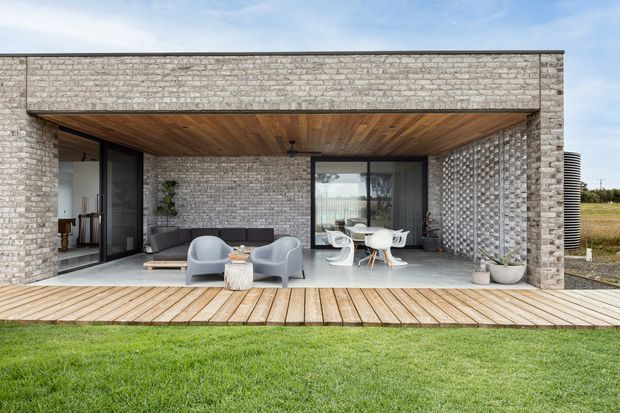 A contemporary single-storey home with a recessed covered outdoor living area. Pale textured brick walls frame large dark-framed glazed openings, while the sheltered ceiling is lined with warm timber boards. A timber deck furnished with lounge chairs and a small dining setting overlooks a lawn, and a perforated masonry screen forms one side of the terrace.