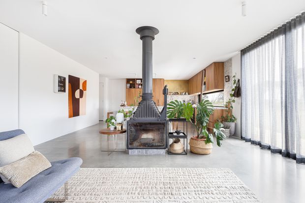 Bright open-plan living area with a freestanding black wood-burning fireplace and exposed flue, pale blue sofa, textured light rug, indoor plants, timber kitchen cabinetry, and full-height sheer curtains. The space has a clean, contemporary feel with warm timber accents and abundant natural light.