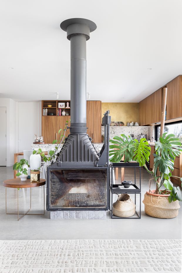 Bright open-plan living area centred on a substantial black freestanding wood-burning fireplace with a tall flue. The room combines warm timber cabinetry and wall panelling, a stone kitchen benchtop, white walls and ceiling, polished concrete flooring, indoor tropical plants, woven baskets, and a low-profile side table. A kitchen and dining area are visible behind the fireplace.