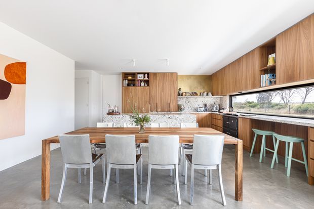 Bright open-plan kitchen and dining area with a long solid timber dining table and six upholstered chairs in the foreground. The kitchen features extensive warm timber cabinetry, a mottled light stone benchtop and splashback, dark appliances, a long horizontal window, and a turquoise bar-stool seating area. Polished concrete flooring and white walls create a clean contemporary interior.