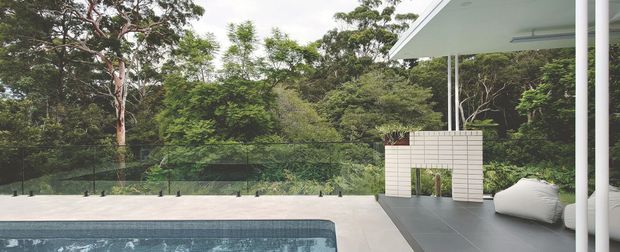 Contemporary outdoor poolside terrace set within dense native greenery. The image shows a rectangular swimming pool, pale paved pool surround, a covered outdoor lounge with dark tiled flooring, a white rendered brick privacy wall, slim white structural posts, and loose white beanbag seating. The setting feels private, calm, and strongly connected to the landscaped bush.