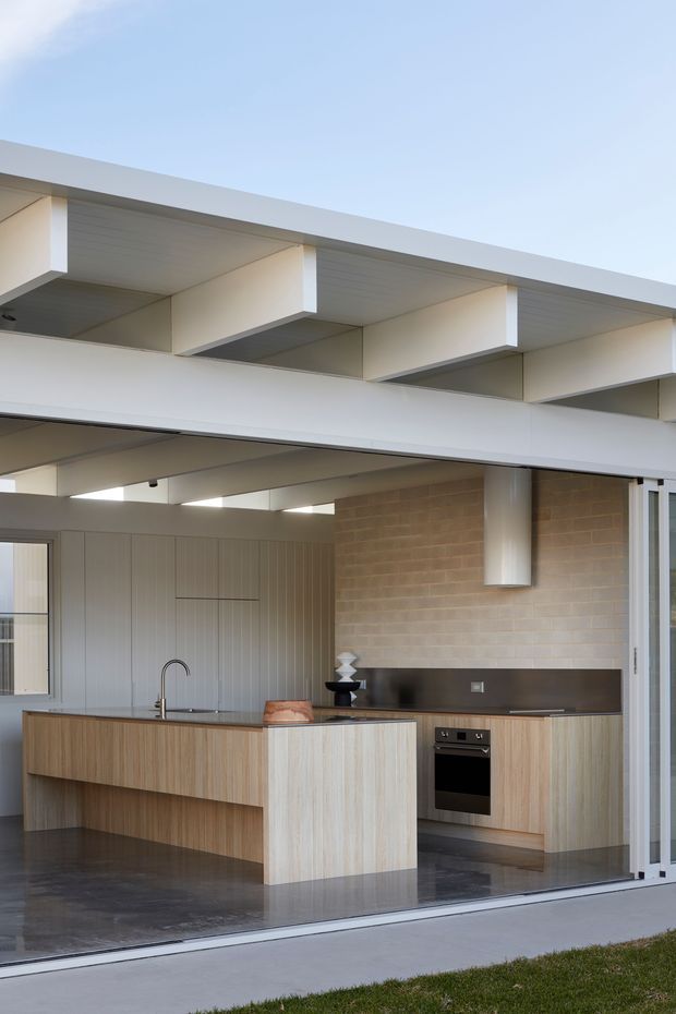 Contemporary open-plan kitchen viewed from outside through large sliding glass doors. The space features a large light timber island with integrated sink, light timber cabinetry, a dark stone-look benchtop and splashback, exposed pale brick walls, polished dark concrete flooring, and a striking open-beam flat-roof ceiling with clerestory-like openings. The palette is restrained and neutral, with strong indoor–outdoor connection.