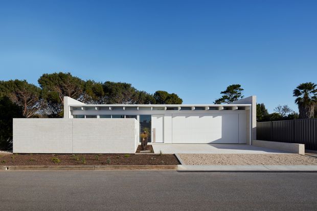 Front elevation of a low, single-storey contemporary residence with a flat roof, white rendered masonry walls, broad glazed entry, and an attached open carport. A light gravel driveway, dark vertical boundary fencing, sparse planting, and mature coastal-looking trees frame the clean minimalist facade.