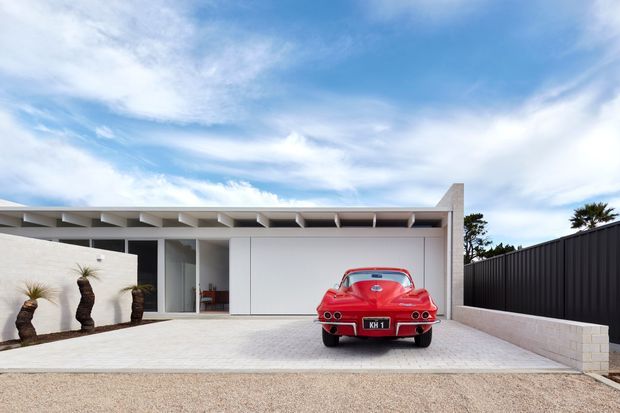 Single-storey contemporary residential facade viewed from the driveway, featuring a broad white rendered garage and entry composition, horizontal concrete or rendered roof-edge detailing, full-height glazing, pale paved forecourt, and a contrasting black vertical-panel boundary fence. A red vintage sports car is parked in front of the garage, with sculptural palms near the entry and a bright blue sky above.