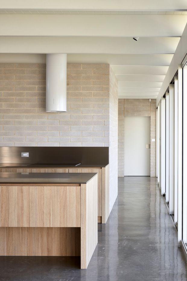 A bright, minimalist kitchen opening onto a corridor, with pale exposed concrete block walls, white-painted structural beams and a cylindrical white rangehood. The kitchen features warm timber-grain cabinetry, dark stone or composite benchtops and splashback, and a polished grey concrete floor. Full-height glazing lines the right side, bringing in abundant natural light.