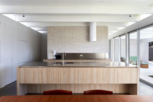 Bright contemporary kitchen viewed from the dining area, featuring a large timber-clad island with a stone benchtop, integrated sink and tap, full-height light brick splashback, stainless-steel rangehood and appliances, extensive glazing, and a white-lined ceiling. Dining chairs and a timber dining table are visible in the foreground, with an outdoor courtyard beyond the glazing.